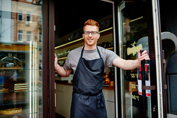 Positive caucasian owner in apron working waiter and opening door of own coffee shop with sweet...