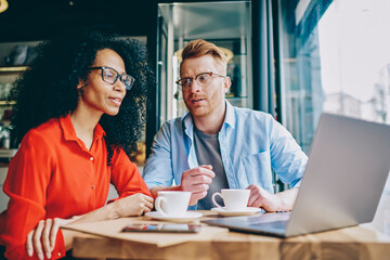 Two multicultural colleagues in eyeglasses watching webinar on laptop device and discussing ideas for development own cafe business.Diverse friends working at computer using wireless internet