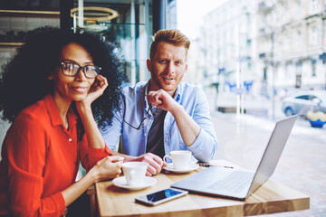 Portrait of positive multicultural male and female friends smiling at camera while spending leisure time togetherness for download movie on laptop computer with good internet connection in cafeteria