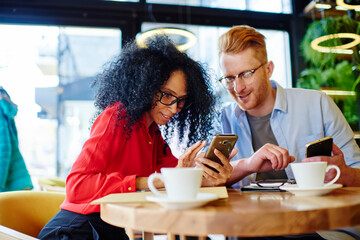 Cheerful adult multiracial couple using smartphones and drinking coffee in modern cafe