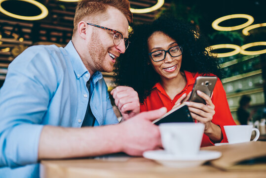 Positive multicultural friends laughing while watching video online on smartphone device using 4G internet connection.Cheerful african american female showing funny news on website caucasian young man