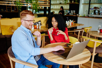 Pensive adult multiethnic entrepreneurs in formal clothes interacting with laptop during business meeting in cozy modern coffee shop