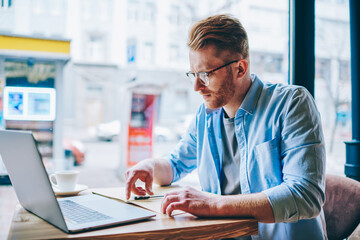 Fototapeta premium Concentrated hipster student reading information and checking data working at modern computer using wireless internet connection in coffee shop.Pensive young man preparing for exam at netbook