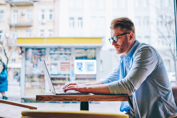 Pensive male graphic designer working on freelance at modern computer using wireless internet in cafe.Concentrated young man in eyewear keyboarding information on digital netbook during distance job