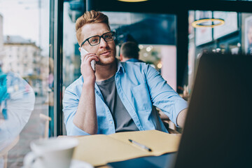 Caucasian young businessman in stylish eyeglasses communicating on smartphone device resting in...