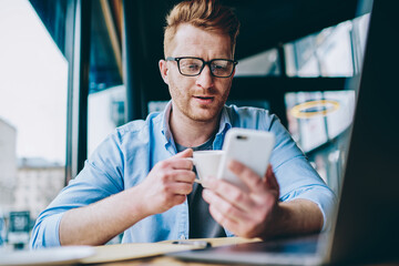 Young man dressed in casual wear drinking tasty coffee while reading news on websites on smartphone using 4G internet.Hipster blogger enjoying caffeine beverage and watching video in social networks