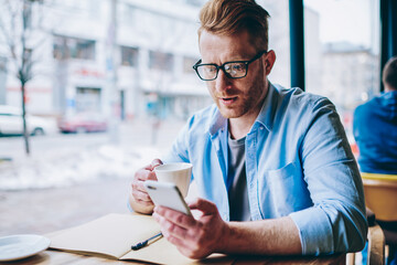 Young businessman checking mail with notification on smartphone device while drinking tasty coffee...