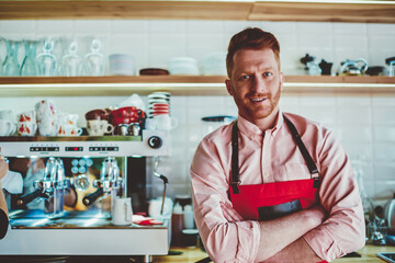 Portrait of successful male barista dressed in uniform working in own cafeteria developing business.Prosperous caucasian waiter with crosses hands standing against bar with modern coffee machine