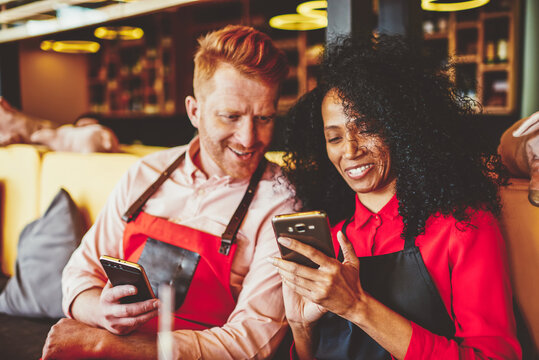 Cheerful diverse friends working together in cafe and discussing funny video in blog on modern smartphone resting during break.Positive waitress showing app on phone smiling male barista in free time