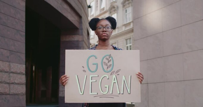 Young woman with Go vegan sign standing at city street and looking to camera. Female afro american student promoting vegetarianism and healthy lifestyle. Concept of eco. Zoom in