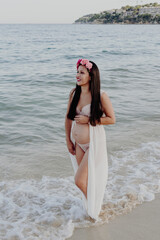 Fototapeta na wymiar Young pregnant Latin woman with flower crown smiling on the seashore. Health during pregnancy, feeling of happiness