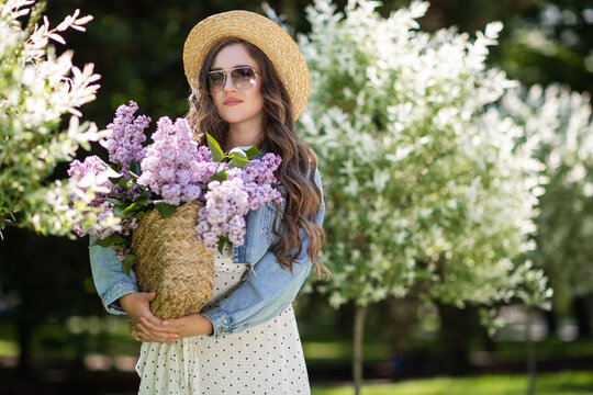 A girl with long hair and straw hat. The girl holds in her hands a wicker basket with flowers. Basket with lilacs. Girl and flowers. Walk with a basket of lilacs in the hands. Floristics.