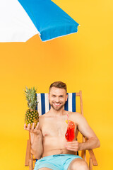  shirtless man sitting on deck chair with cocktail and pineapple near beach umbrella on yellow