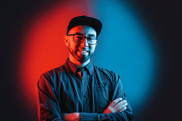 Neon lights. Portrait of a man in a shirt, glasses and cap, with a beard, posing with his arms...