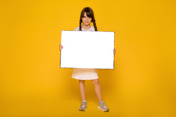 Beautiful School girl holding white sheet. Cute kid with paper. Yellow background. Copy spase....