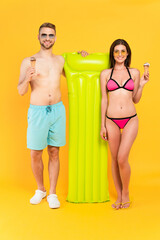  muscular man and cheerful woman in swimwear and sunglasses holding ice cream while standing near inflatable mattress on yellow