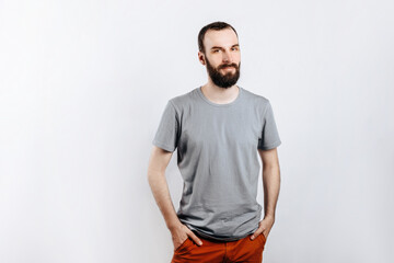 A handsome brutal brunette man with a beard in a gray shirt smiles and looks at the camera, holds his hand in his pockets. Place for advertising on a gray background.