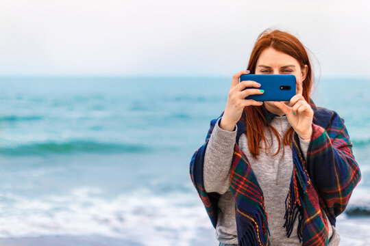 Beautiful girl with long brown hair takes pictures on smartphone on the beach near the ocean, vacation concept