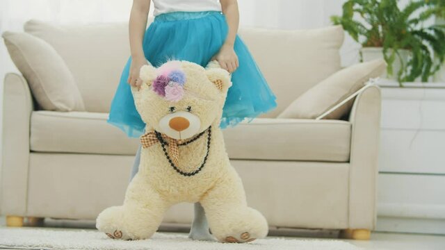 Cropped little girl in blue skirt standing, holding a plush teddy bear in necklace and funny headgear.