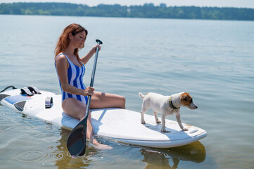 A woman is riding a sup surfboard with a dog on the lake. The girl goes in for water sports with her pet.