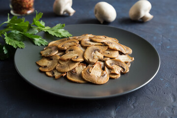 Slices of fried mushrooms on a black plate with of parsley leaves and several whole champignons. Close up on a dark blue background.
