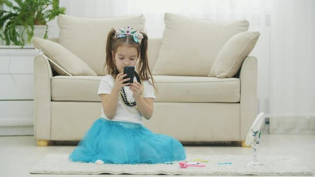 Beautiful little kid wearing crown, dress, necklace, and sitting on the carpet with phone in her hands.
