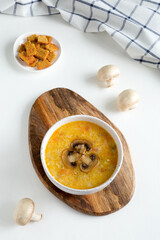Champignon soup in white bowl on wooden stand. Whole champignons, crackers flat lay on a concrete white background. Top of the soup is decorated with slices of sliced mushroom.