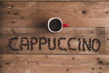Top view composition of red mug with roasted aromatic coffee placed on wooden table above inscription cappuccino made with coffee beans