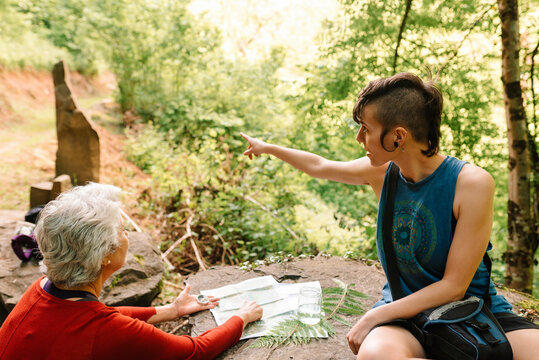 Side view of young and senior female travelers sitting on stone in woods and discussing location while orientating on map and compass