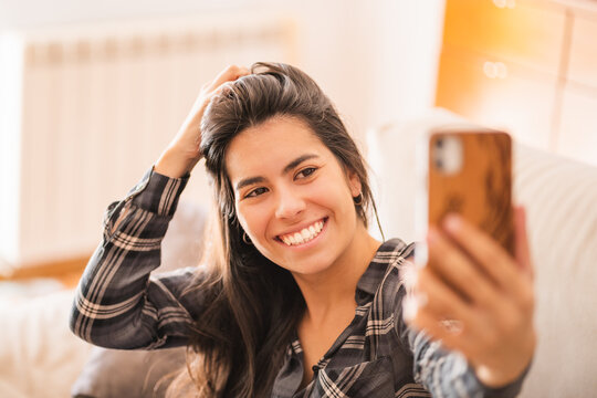 High angle of optimistic young female in casual wear smiling happily while sitting on sofa and taking selfie with mobile phone during free time at home