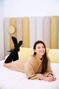Charming female in oversize sweater and stockings lying on soft bed in modern bedroom and cheerfully looking at camera