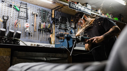 Side view of bearded male master wearing protective glasses standing in shabby workshop and cutting metal detail with grinder tool