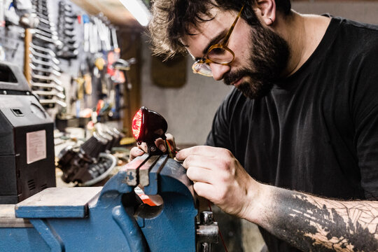 Side view of concentrated male mechanic with tattoos using vise and repairing red back lamp of motorbike while working in workshop