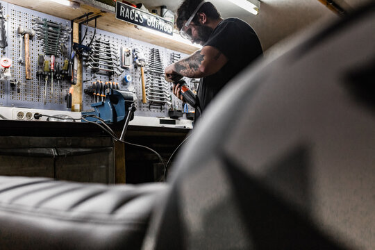 Side view of bearded male master wearing protective glasses standing in shabby workshop and cutting metal detail with grinder tool