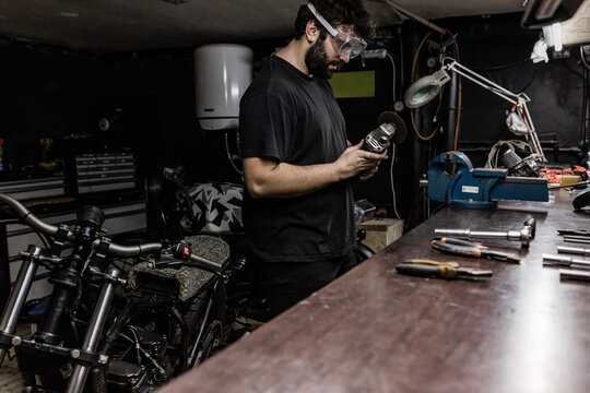 Side view of bearded male master wearing protective glasses standing in shabby workshop and cutting metal detail with grinder tool