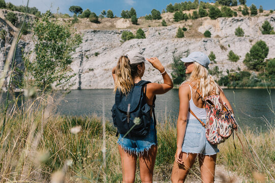 Back view of carefree women in shorts standing on grass and admiring spectacular view of lake and hills on sunny day