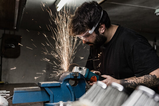 Side view of bearded male master wearing protective glasses standing in shabby workshop and cutting metal detail with grinder tool