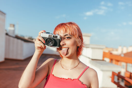 Naughty female millennial with piercing and pink hair taking pictures on vintage photo camera while showing tongue and looking at camera