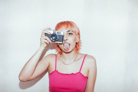 Naughty female millennial with piercing and pink hair taking pictures on vintage photo camera while showing tongue looking at camera on a white wall background