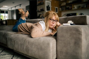 Side view of pensive female in casual wear lying on sofa at home and enjoying interesting novel while relaxing during weekend