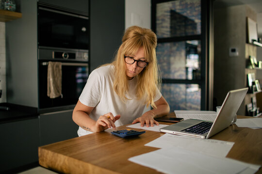 Focused female bookkeeper in casual outfit sitting at table in kitchen and counting bills while doing paperwork using calculator