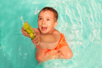 Fototapeta na wymiar Little toddler boy sitting in swimming pool with funny face expression and shooting (or aiming) up with green water gun. Summer weekend, vacation and outdoors activitiy for kids concept