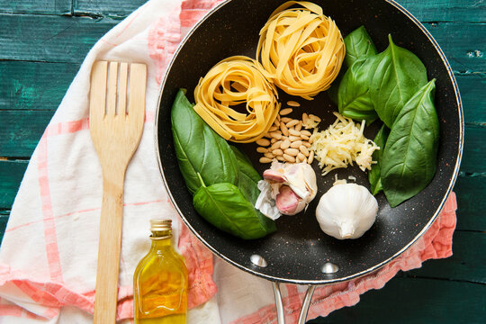 Top view of bottle of olive oil near frying pan with raw tagliatelle rolls surrounded by heads of garlic with green basil leaves and crispy pine nuts