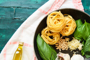 Top view of bottle of olive oil near frying pan with raw tagliatelle rolls surrounded by heads of garlic with green basil leaves and crispy pine nuts