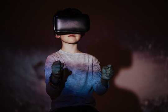 Cute boy wearing VR glasses and exploring virtual reality while standing under colorful projection