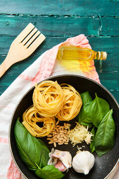 Top view of bottle of olive oil near frying pan with raw tagliatelle rolls surrounded by heads of garlic with green basil leaves and crispy pine nuts
