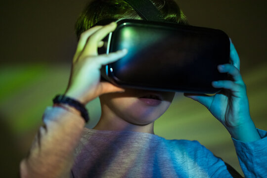 Cute boy wearing VR glasses and exploring virtual reality while standing under colorful projection
