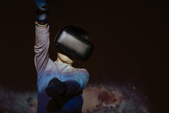 Cute boy wearing VR glasses and exploring virtual reality while standing under colorful projection
