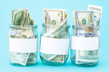 Three Piggy Bank from a Glass Jar for Life. New dollars sticking out of piggy banks on a blue background. The banks have white sheets for inscriptions. Mockup. Copy space. Economy concept