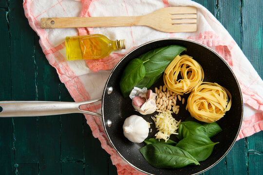 Top view of frying pen with uncooked pasta rolls near fresh basil leaves and heads of garlic with pine nuts and bottle of olive oil near wooden spatula on towel